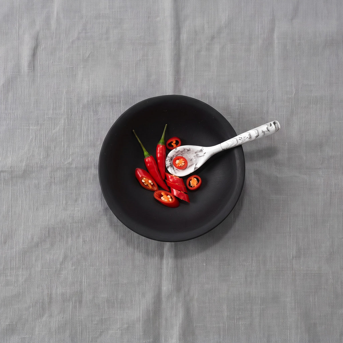 POLKA LUKA matte black resin relish bowl with a white marble-effect spoon containing sliced red chili peppers