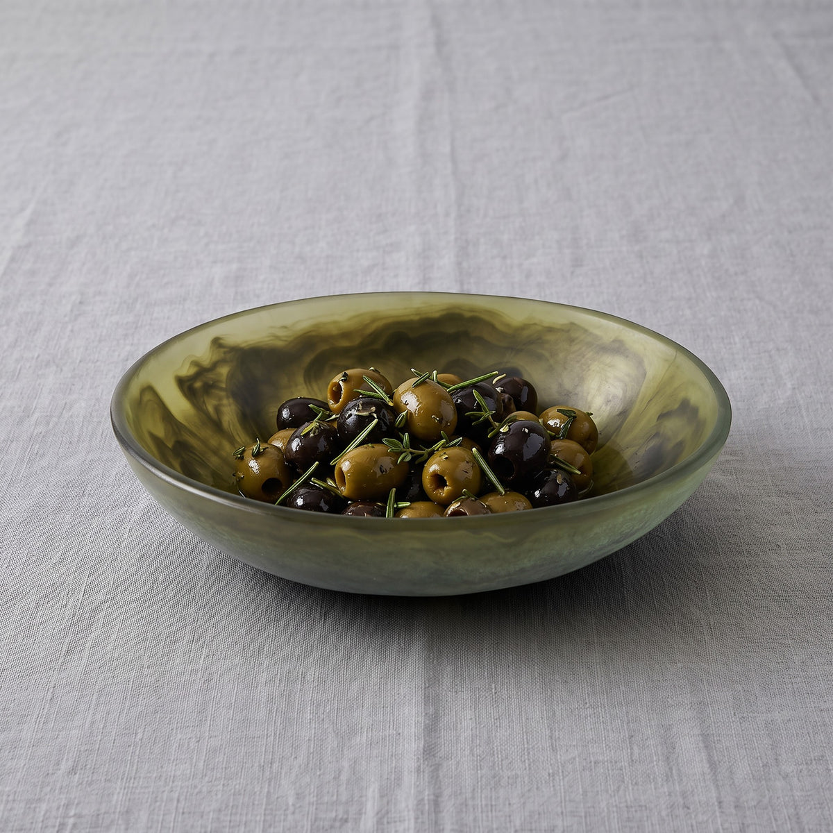 POLKA LUKA small oval resin bowl in a marbled olive green finish, filled with olives on a grey linen tablecloth.